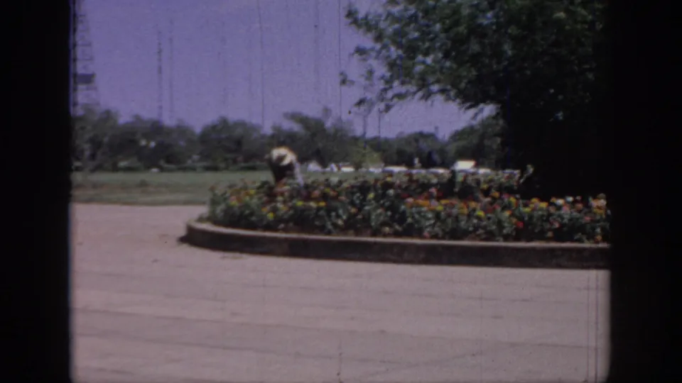 1967 Oklahoma 8mm frame showing a distant person in a light hat working near a circular flower bed, with trees and a utility tower behind.