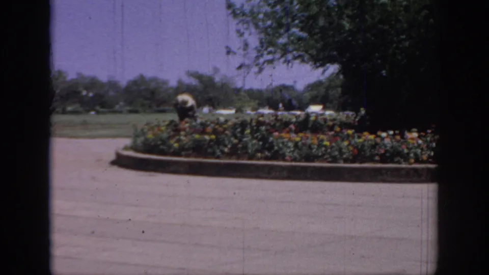 Distant gardener figure appears behind a circular flower bed in 1967 Oklahoma footage, with trees, lawn, and pale sky in the background.
