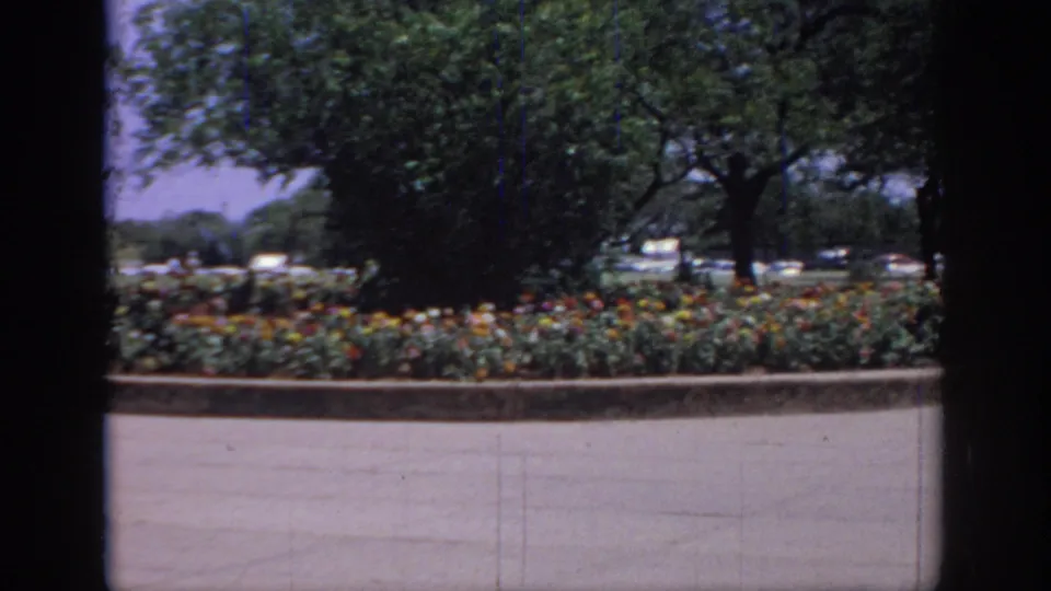 1967 Oklahoma 8mm frame showing colorful flower beds, trees, pavement, and dark window framing in the foreground.
