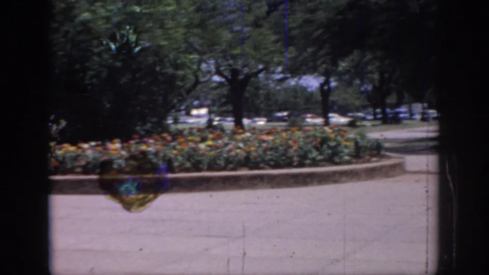 1967 Oklahoma home-movie frame with a curved flower bed, trees, pavement, and dark vertical framing across the garden view.