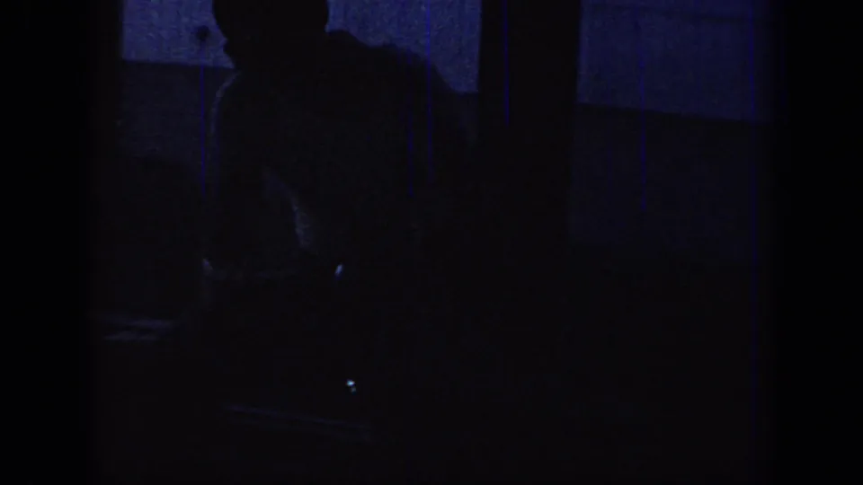 Underexposed 1967 Oklahoma 8mm interior frame with a dark seated silhouette, blue-toned wall, and vertical framing.