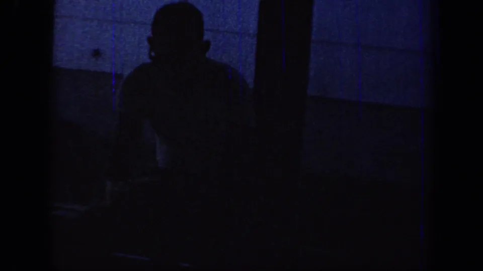 Low-light 1967 Oklahoma home-movie frame showing a dark human-like silhouette against a blue-toned interior surface.