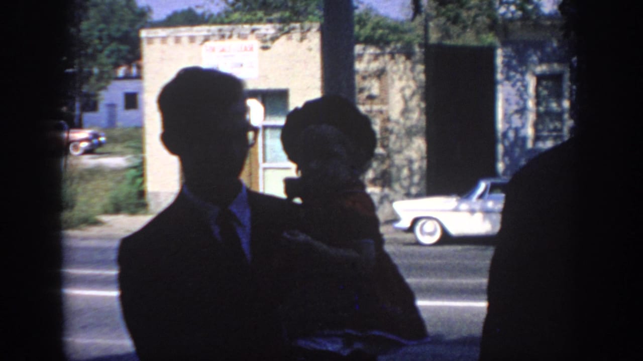 1967 Oklahoma home-movie frame with a silhouetted man holding a small child near a sunlit street and mid-century cars.