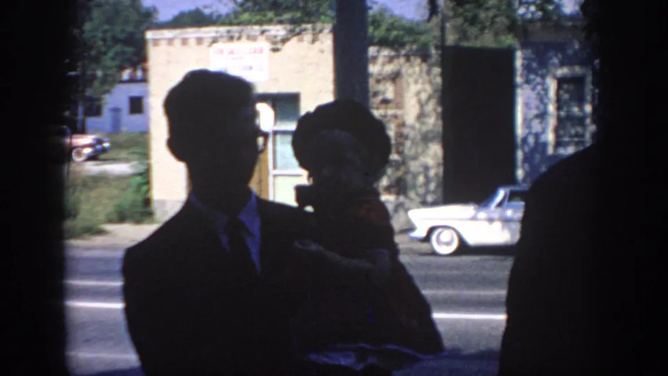 1967 Oklahoma home-movie frame with a silhouetted man holding a small child near a sunlit street and mid-century cars.