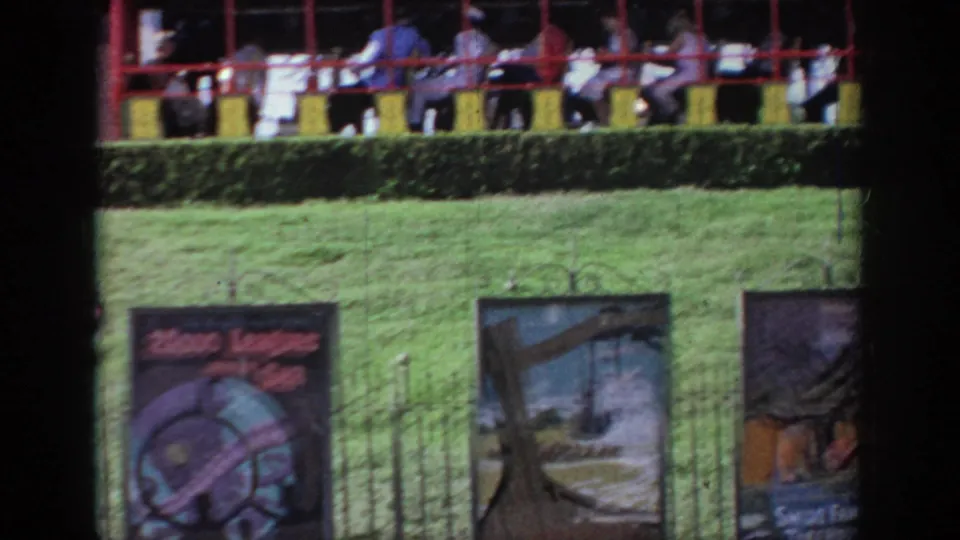1960s home-movie frame of a small passenger train moving behind a hedge, with attraction signs on a green lawn.