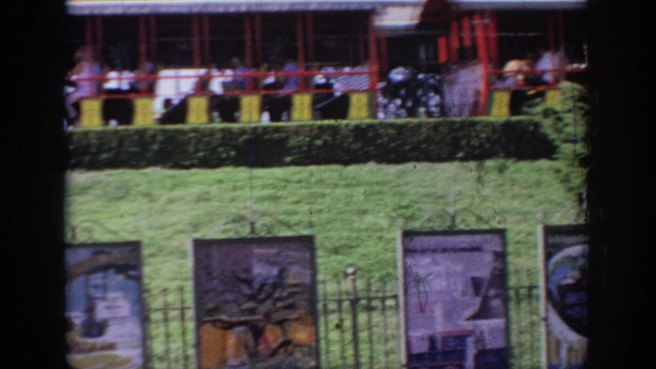 1967 Oklahoma home-movie frame showing passengers in red-and-yellow elevated ride cars above attraction banners and a fence.