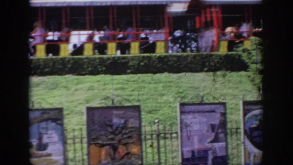 1967 Oklahoma home-movie frame showing passengers in red-and-yellow elevated ride cars above attraction banners and a fence.