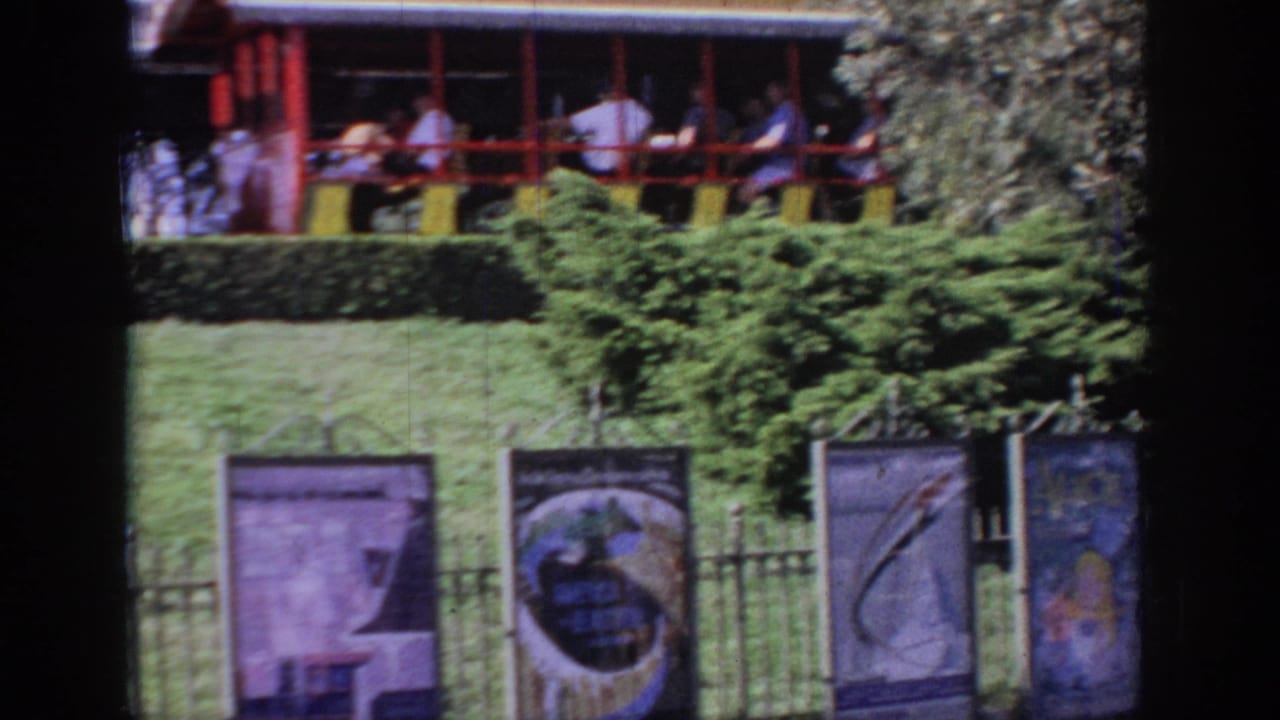 Passengers ride past shrubs and attraction banners behind a fence.