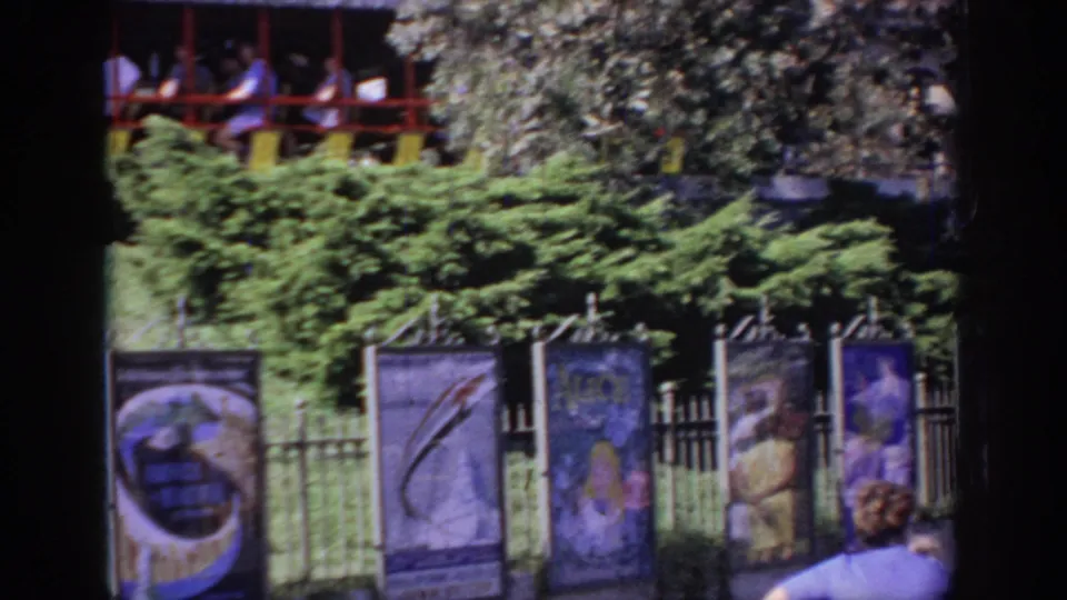 1967 Oklahoma amusement-park view with banner panels, a fence, shrubs, and a partial elevated ride car at the top of frame.