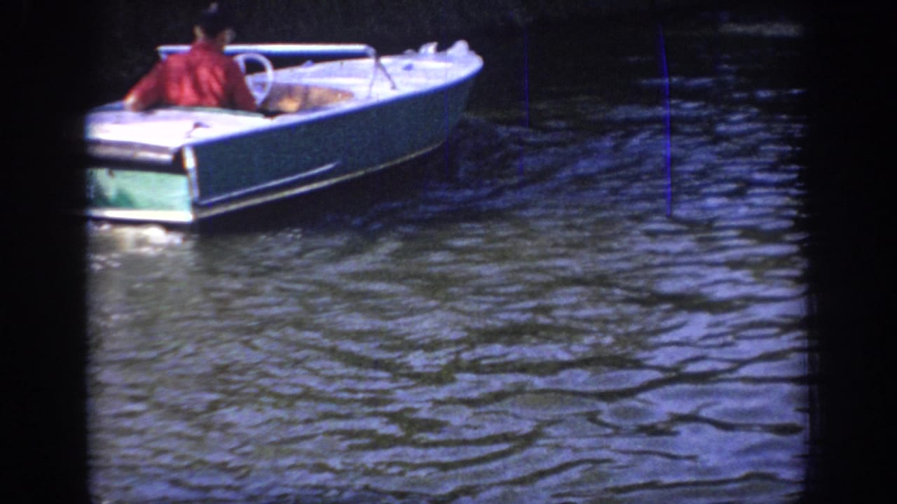 1967 Oklahoma 8mm frame showing a man from behind in a white-and-green motorboat on a shaded river.