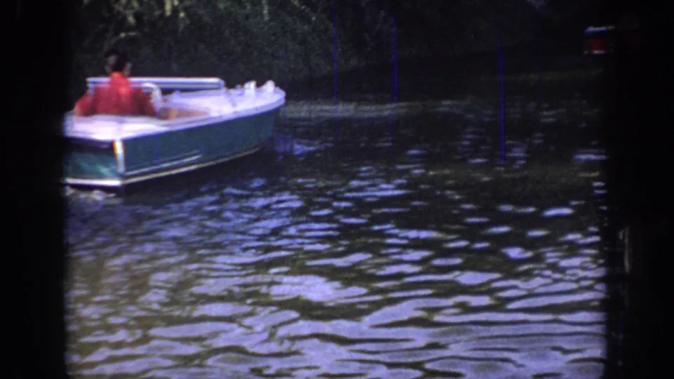 1967 Oklahoma 8mm still of a man from behind in a white motorboat crossing dark water beside dense trees.