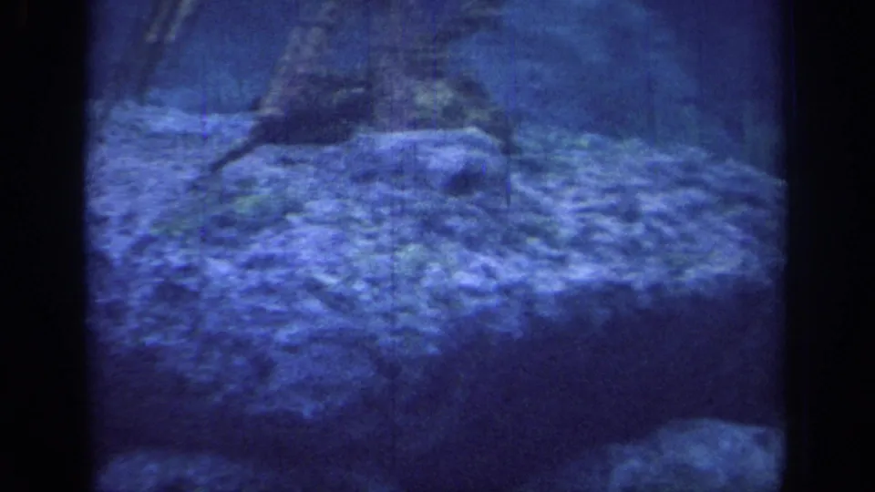 Blue-shifted 1967 8mm frame from Oklahoma showing a rocky ledge or peak area with dark tree forms and film gate edges.
