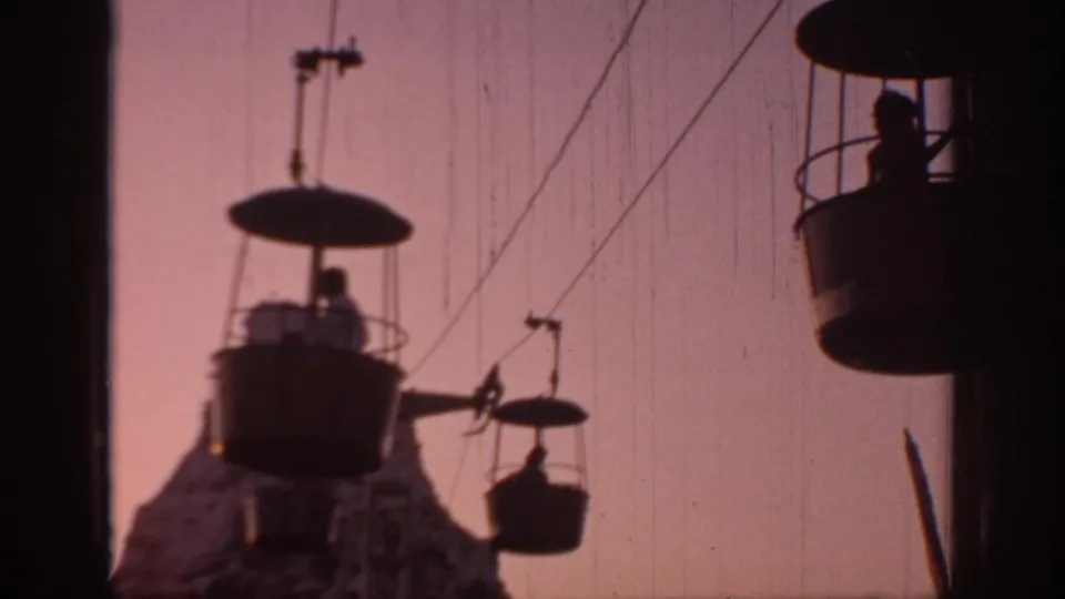 1967 Oklahoma 8mm frame showing several gondola cabins on overhead cables, with small silhouetted riders and a rocky peak below.
