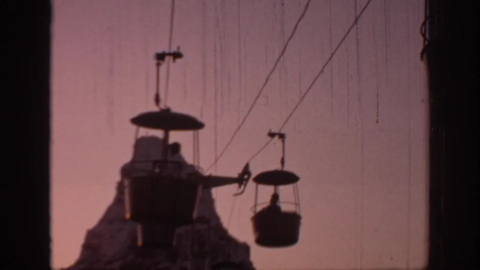 A wider 1967 Oklahoma home-movie frame of gondola cabins moving on cables above a rocky silhouette at dusk, with riders visible as dark figures.