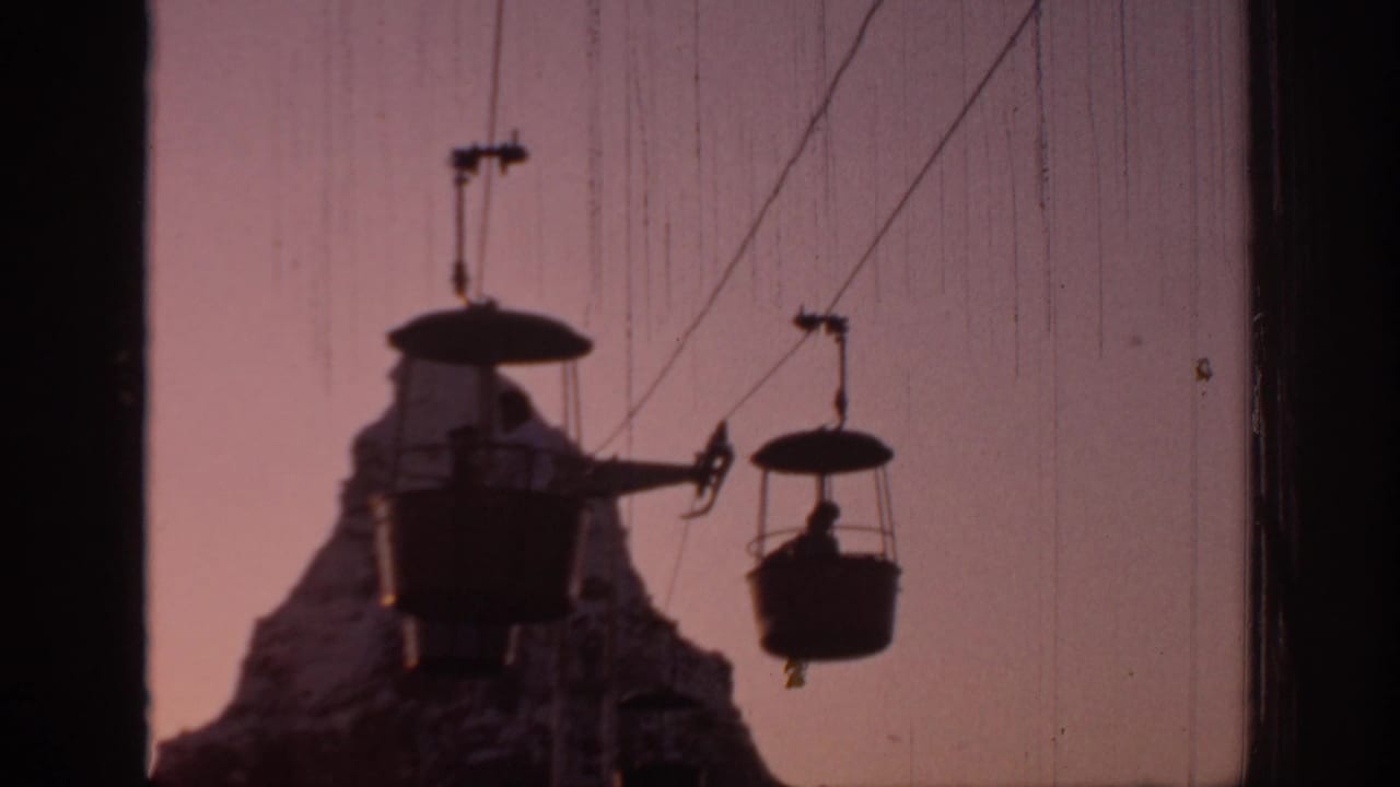 Suspended amusement-park cable cars pass in front of a mountain-shaped structure at sunset.