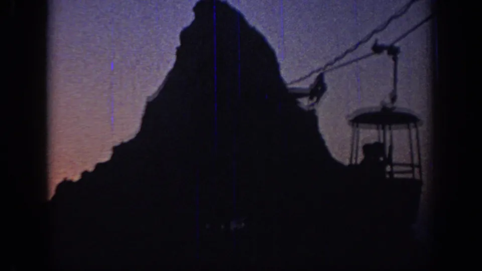 1967 Oklahoma frame showing a silhouetted rider in a suspended cable car beside a mountain-shaped attraction at sunset.
