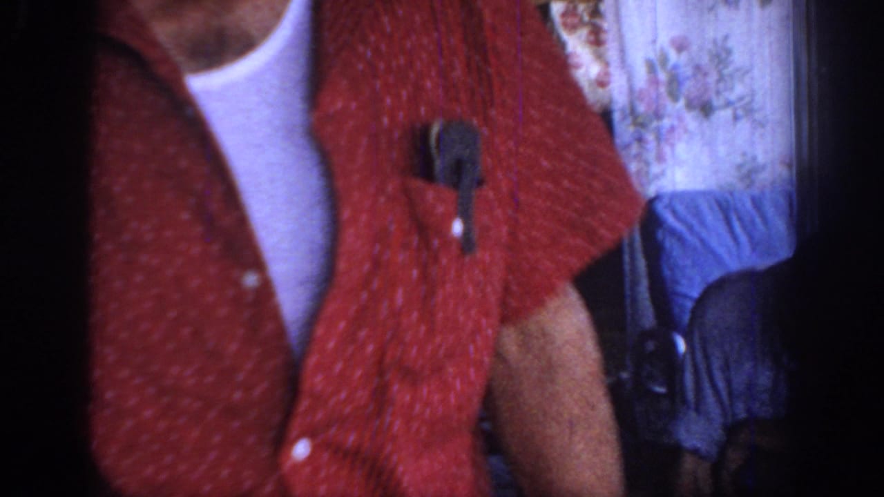 1967 Oklahoma home-movie frame showing a close cropped adult torso in a red patterned shirt with a white undershirt in a dim interior.