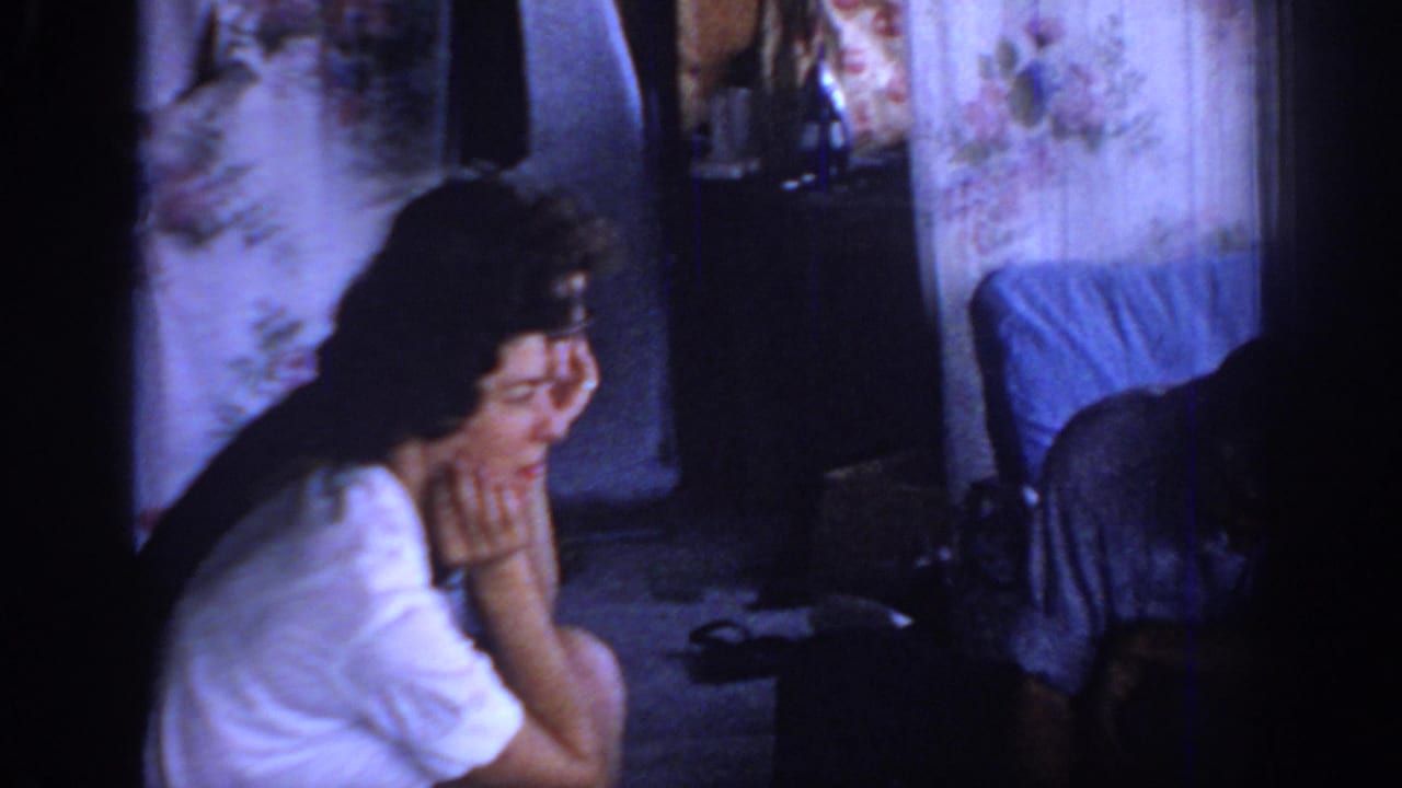1967 Oklahoma home-movie frame of a woman in a white blouse seated in a dim private room with floral curtains and covered furniture.