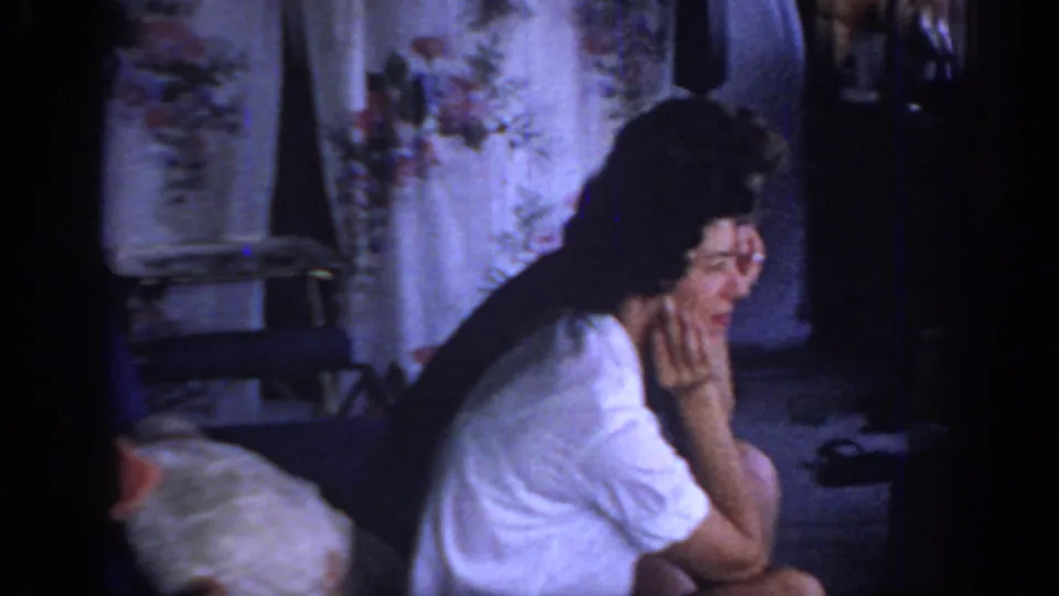 1967 Oklahoma home-movie frame showing a seated woman in a white top inside a private room with floral curtains and low light.