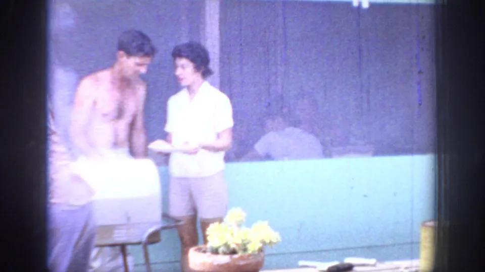 1967 Oklahoma home-movie frame of an outdoor patio scene with a shirtless man, woman holding a plate, grill, wall, and potted flowers.