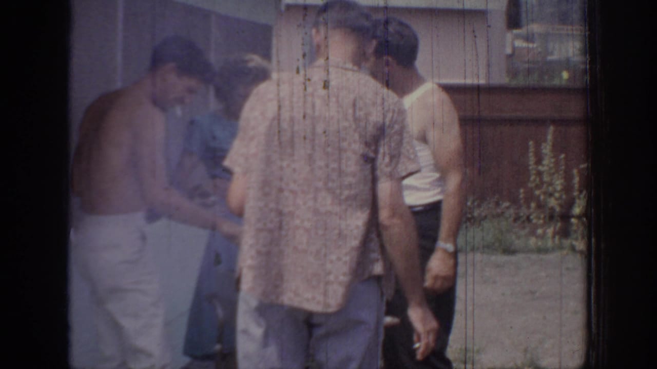 1967 Oklahoma 8mm frame showing adults gathered outdoors near a house, fence and plants during a casual social moment.