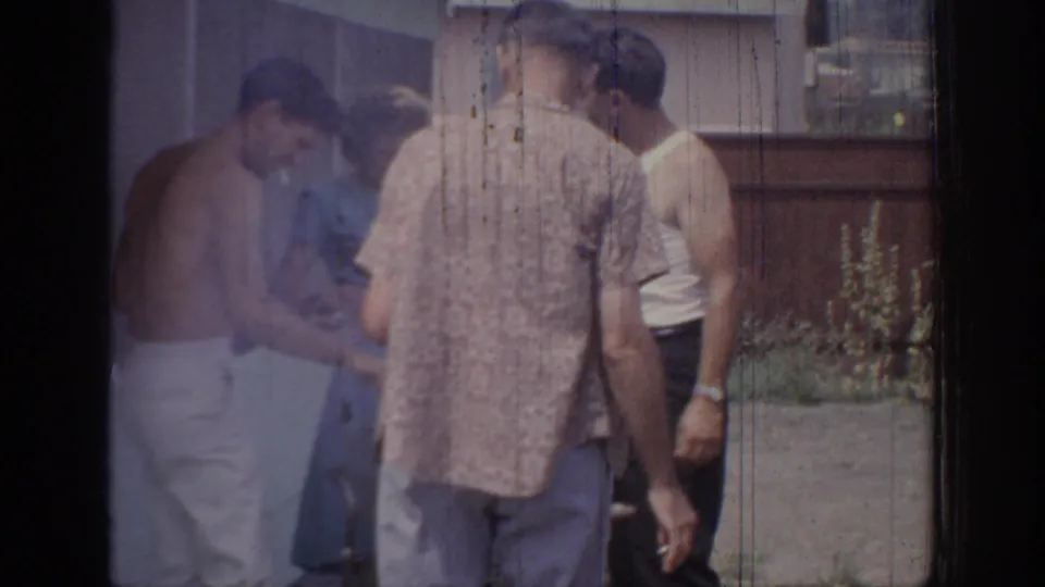 1967 Oklahoma 8mm frame showing adults gathered outdoors near a house, fence and plants during a casual social moment.