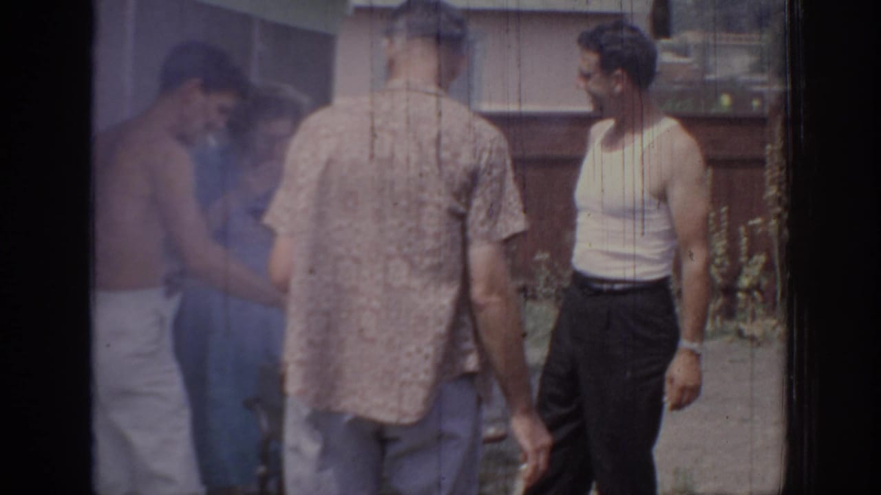 1967 Oklahoma home-movie frame of adult men standing in conversation beside a wooden fence and house exterior.