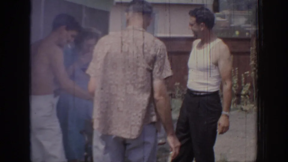1967 Oklahoma home-movie frame of adult men standing in conversation beside a wooden fence and house exterior.