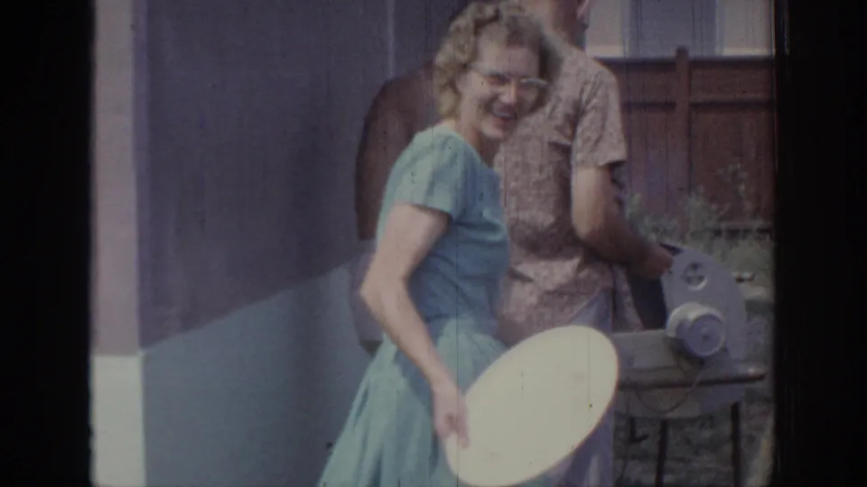 In 1967 Oklahoma home-movie footage, a woman in a light blue dress smiles beside adults near a backyard grill area.