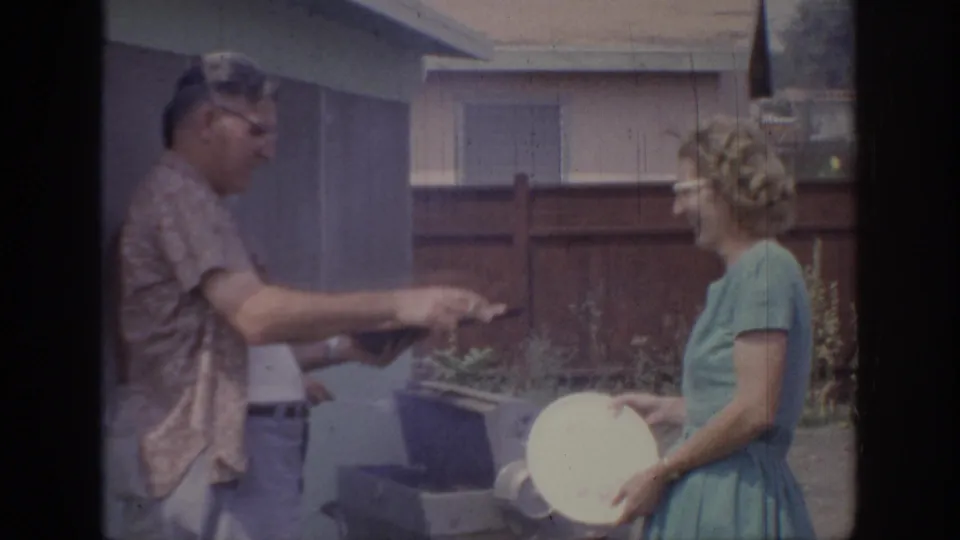A man in glasses gestures near a woman holding a white plate beside a small backyard grill in 1967 Oklahoma footage.