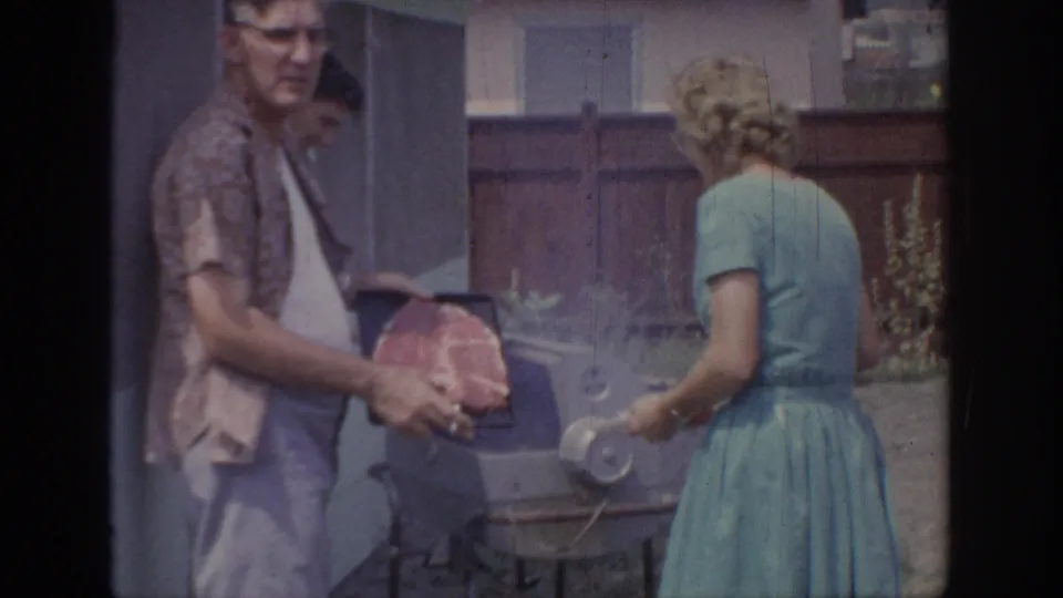 A man holds raw meat beside a small grill while a woman in a blue dress holds a plate in the Oklahoma backyard scene.