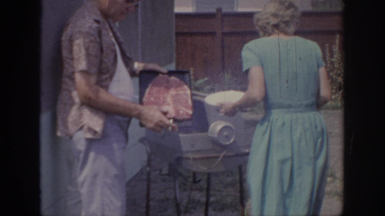 1967 Oklahoma home-movie frame of a man holding steak near a backyard grill beside a woman in a teal dress with a plate.