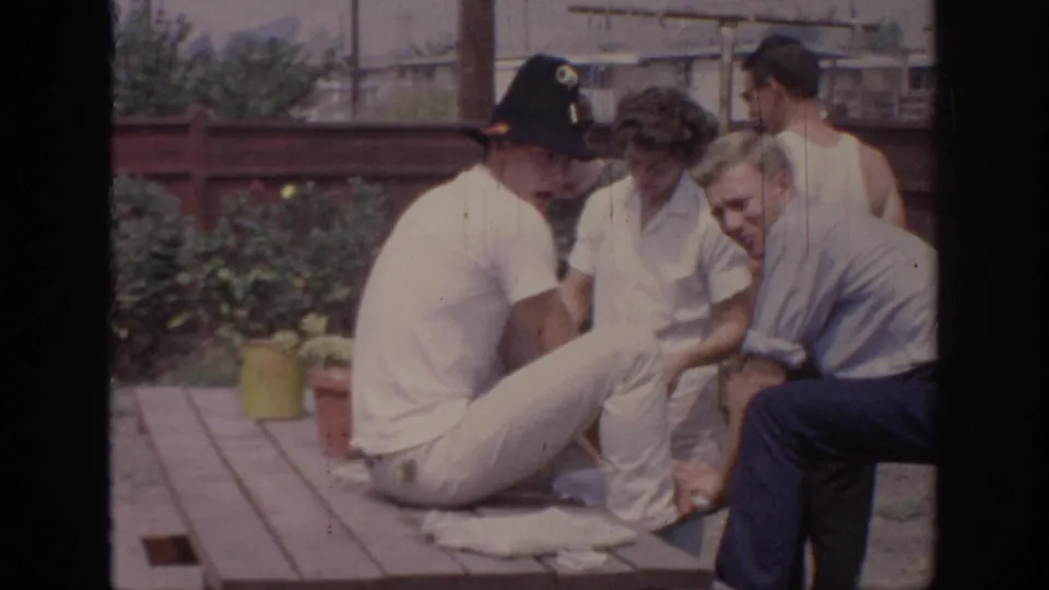 Home-movie frame from a 1967 Oklahoma backyard party showing adults sitting together at a wooden table.