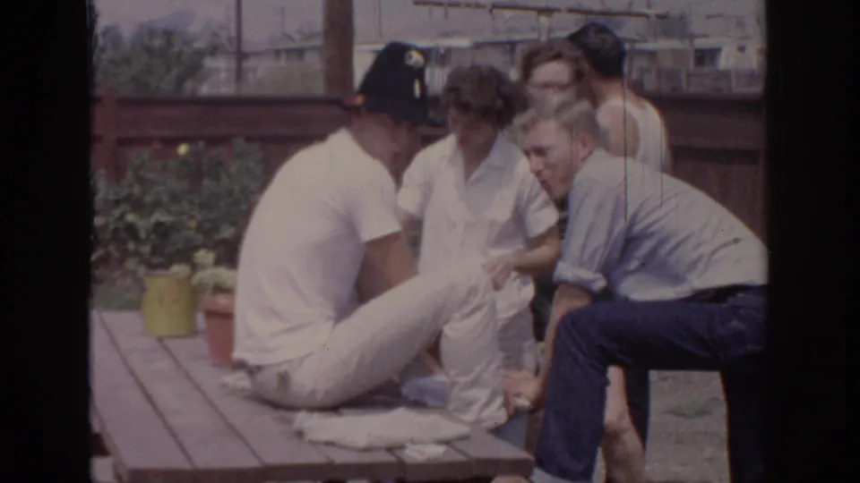 1967 Oklahoma home-movie frame of adults socializing around a wooden picnic table in a residential backyard.
