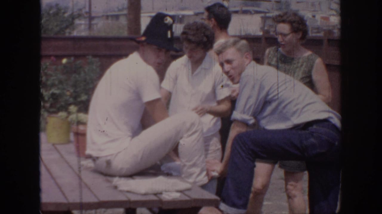 1967 Oklahoma 8mm frame showing adults gathered around a seated man in white trousers and black hat at a backyard picnic table.