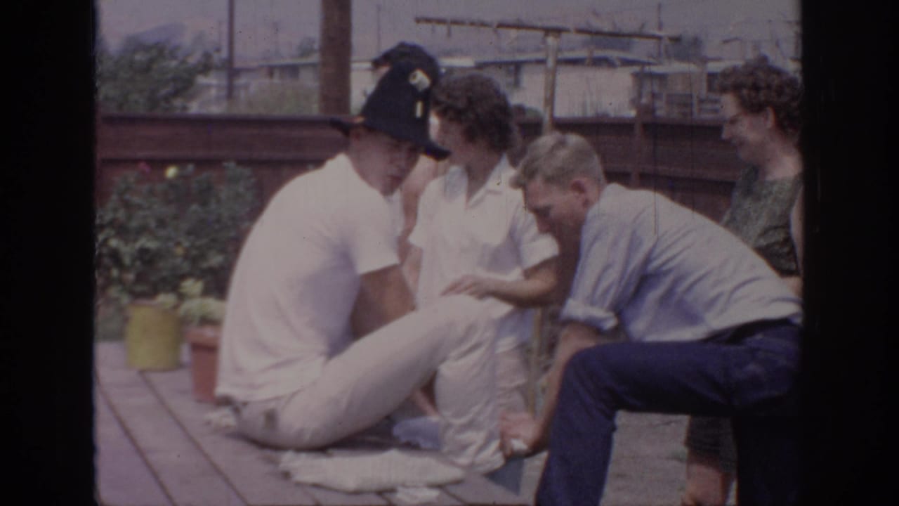 A seated man in a black hat turns near friends beside a picnic table.
