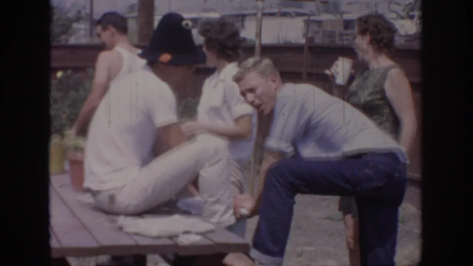 1967 Oklahoma home-movie frame with adults moving around a backyard picnic table, cups visible among the group.