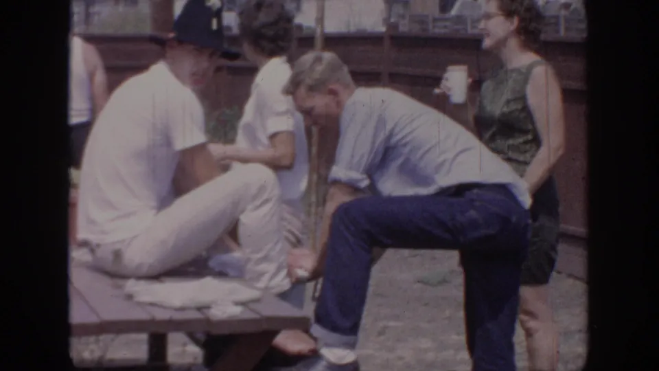 Adults at a 1967 Oklahoma backyard gathering stand and sit close together, with a man in blue leaning beside the picnic table.