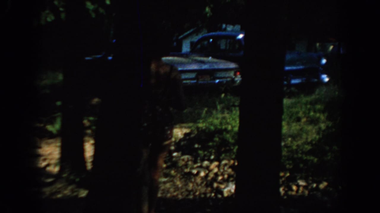 1967 Oklahoma 8mm frame showing a blue sedan glimpsed through dark tree trunks in a wooded outdoor area.
