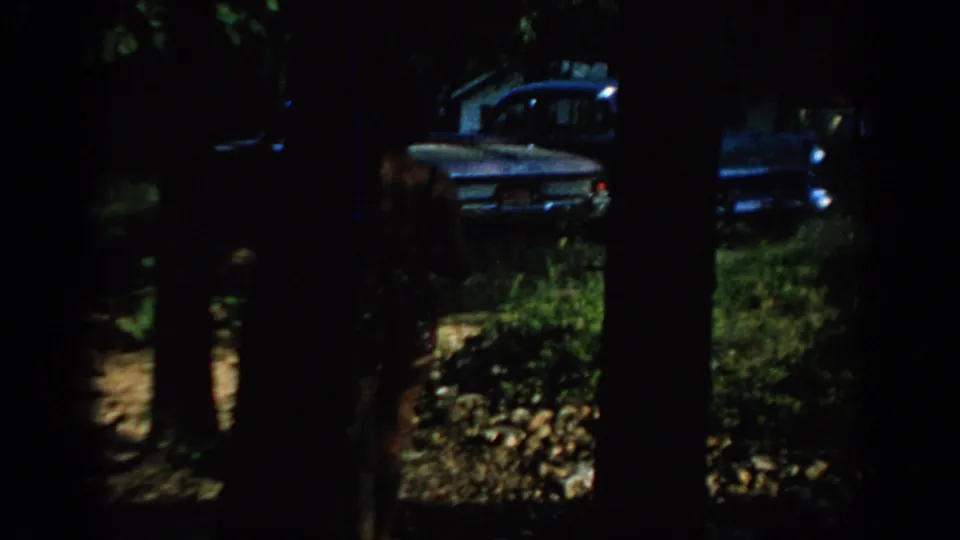 1967 Oklahoma 8mm frame showing a blue sedan glimpsed through dark tree trunks in a wooded outdoor area.