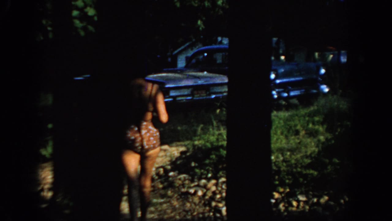 Woman in a patterned bikini walks toward a blue sedan through trees.
