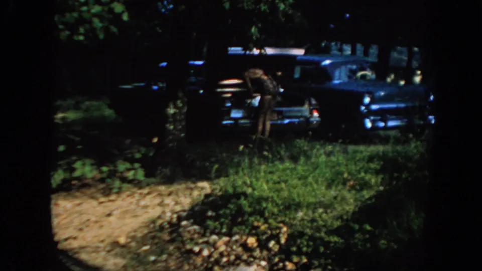 1967 Oklahoma 8mm frame showing a woman near the rear of a blue sedan, seen through dark tree trunks.