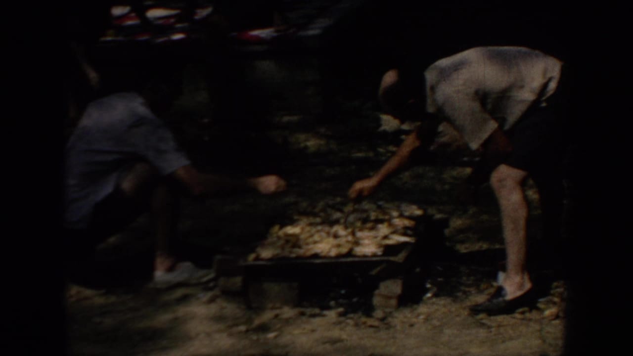 1967 Oklahoma home-movie frame showing a man bending over a low grill while another person sits nearby in a wooded park.