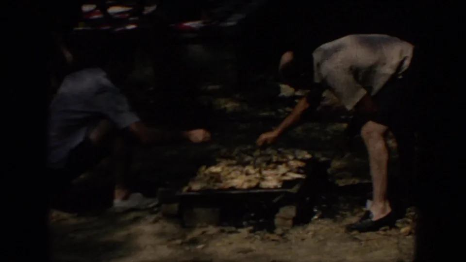 1967 Oklahoma home-movie frame showing a man bending over a low grill while another person sits nearby in a wooded park.
