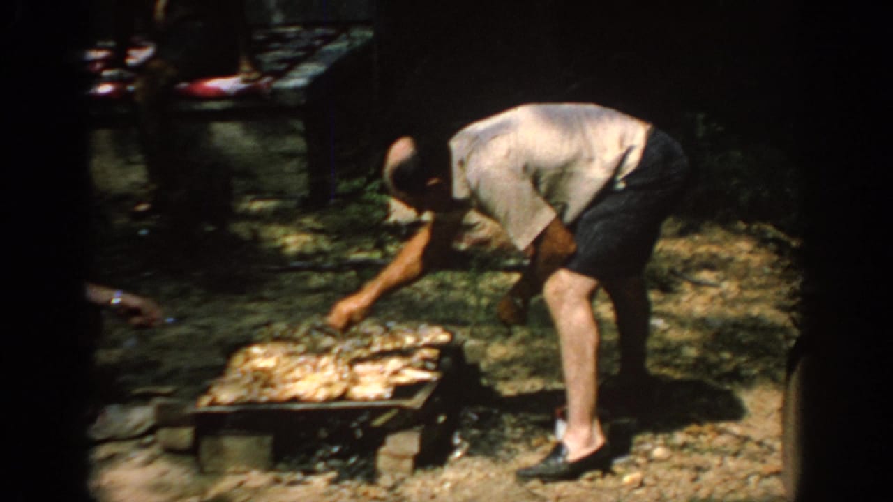 1967 Oklahoma frame of a man in a light shirt and dark shorts tending a low barbecue grill under trees.