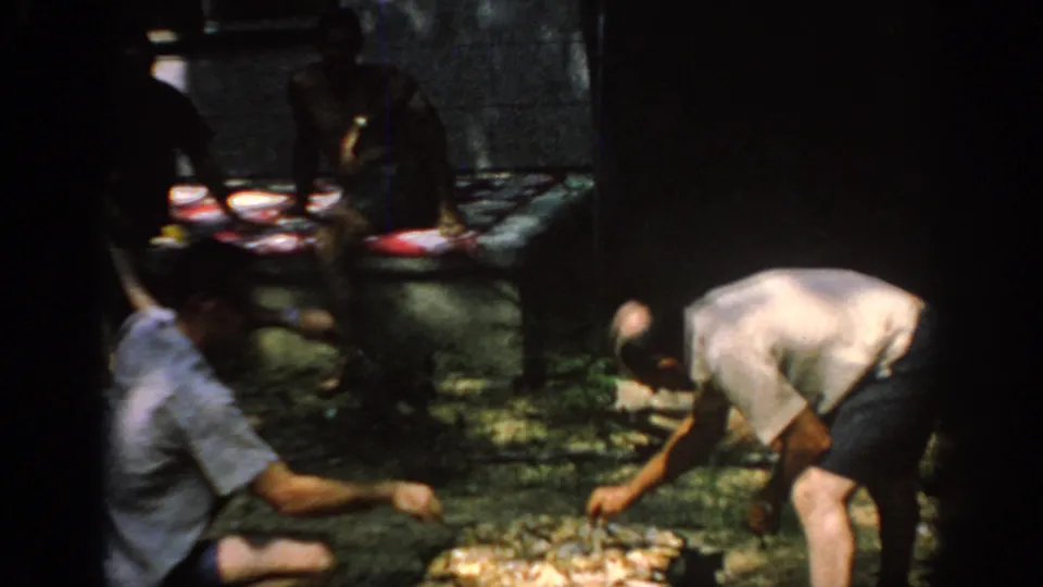 1967 Oklahoma home-movie frame with a man cooking at a low grill and companions seated near picnic plates behind him.