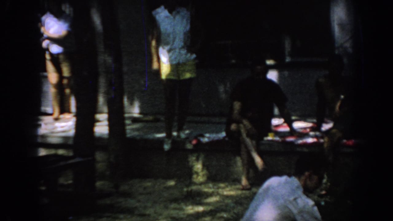 1967 Oklahoma 8mm frame showing adults near a shaded porch, with seated figures and tree trunks obscuring parts of the view.