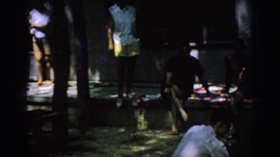 1967 Oklahoma 8mm frame showing adults near a shaded porch, with seated figures and tree trunks obscuring parts of the view.