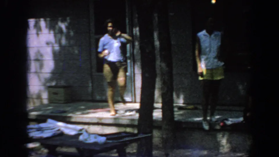 1967 Oklahoma 8mm frame showing adults around porch steps, with tree trunks, shadow, and scattered cloth in the foreground.