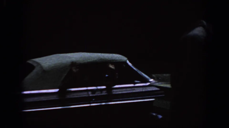 Shadowed 8mm frame showing part of a parked car before the backyard laundry scene begins.